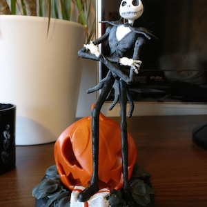 Può includere: Una statuetta stampata in 3D in bianco e nero di Jack Skellington de The Nightmare Before Christmas in piedi su una zucca con una scatola regalo. La statuetta è in posa con le braccia incrociate e un leggero sorriso.