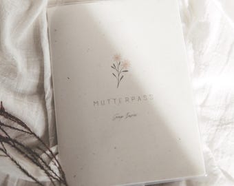 May include: A rectangular off-white booklet with a speckled texture, featuring a small illustration of two pink flowers and the text MUTTERPASS printed in black, with additional cursive text below the main title, encased in a clear protective cover.