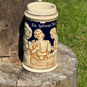 May include: A ceramic beer stein with a cream-colored lid and base, and a dark blue midsection. The midsection features a relief of three figures and the text "Ein heiteres Trio". The stein is sitting on a wooden stump.