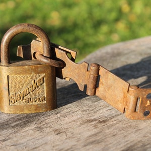 May include: A vintage brass padlock and a rusty metal hinge. The padlock is marked "Slaymaker SUPER" within a rectangular frame. The metal shows signs of age and wear, with a weathered appearance. The set is resting on a wooden surface.