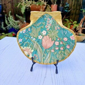 May include: A decorative ceramic seashell dish with a floral design in shades of green, pink, and gold. The dish is on a black metal stand.