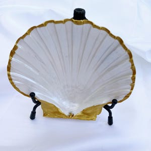 May include: A white seashell with a gold rim, displayed on a black metal stand. The seashell is a decorative object.