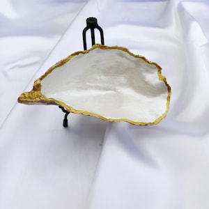 May include: White ceramic oyster shell dish with a gold rim, displayed on a black metal stand.