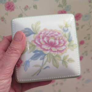 May include: A square porcelain trinket box with a floral design. The box features a pink and yellow flower with green leaves on a white background. The box has a green trim and a beaded edge.