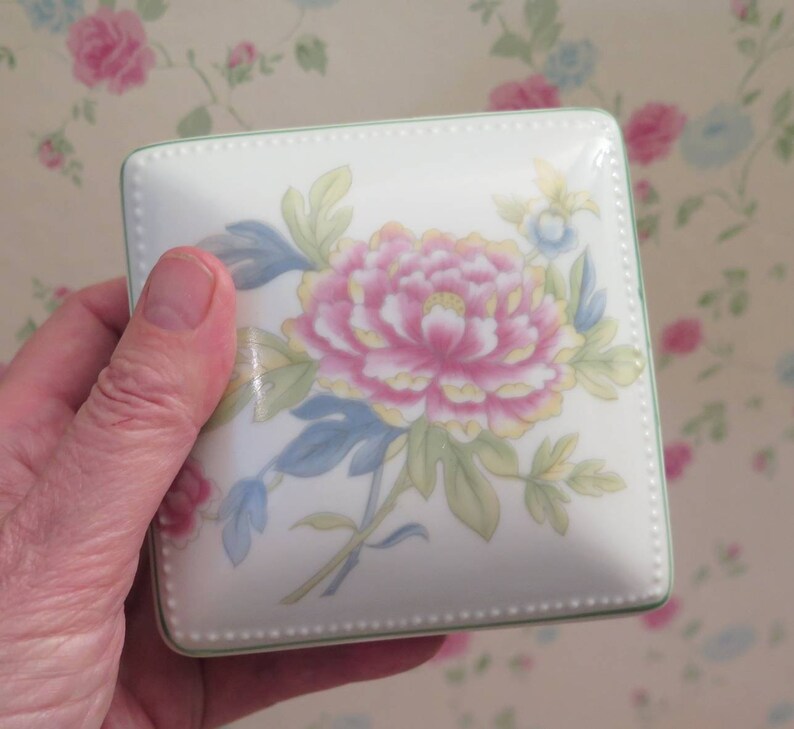 May include: A square porcelain trinket box with a floral design. The box features a pink and yellow flower with green leaves on a white background. The box has a green trim and a beaded edge.
