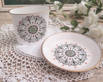 May include: A white oval-shaped ceramic cup and saucer set with a green, red, and black geometric design. The cup and saucer are on a white crocheted doily.