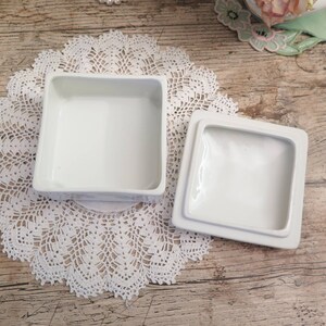 May include: A white porcelain box with a square lid. The box is open and the lid is sitting next to it. The box is on a white doily.