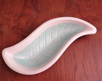 May include: A ceramic leaf-shaped dish with a light pink border and a pale green interior. The interior features a detailed leaf vein pattern. The dish is placed on a dark wood surface.