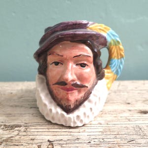 May include: A ceramic mug shaped like a man's head, featuring a purple hat, a white ruffled collar, and a handle with yellow and blue stripes. The mug has a painted face with a mustache and beard.