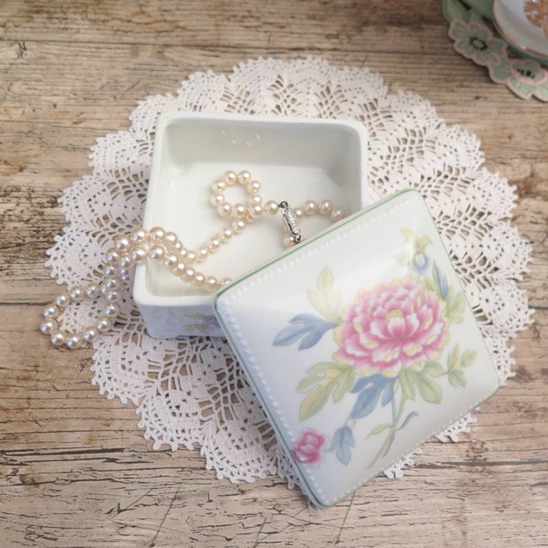 May include: A white porcelain trinket box with a floral design. The lid features a pink and yellow flower with green leaves. The box is open and a pearl necklace is inside.