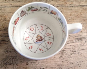 May include: A white ceramic teacup featuring a central illustration of a sailing ship surrounded by a compass-like star design with red astrological symbols. The inner rim displays a series of small colourful icons including a snake and fruit.