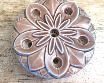 May include: A decorative, round, ceramic object with a floral design. The object is a muted mauve colour with a glossy finish and features a central hole surrounded by six smaller holes. The design is raised and detailed, with a geometric border.
