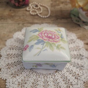 May include: A white porcelain square trinket box with a floral design. The box has a green trim and a delicate floral pattern with a pink flower in the centre.