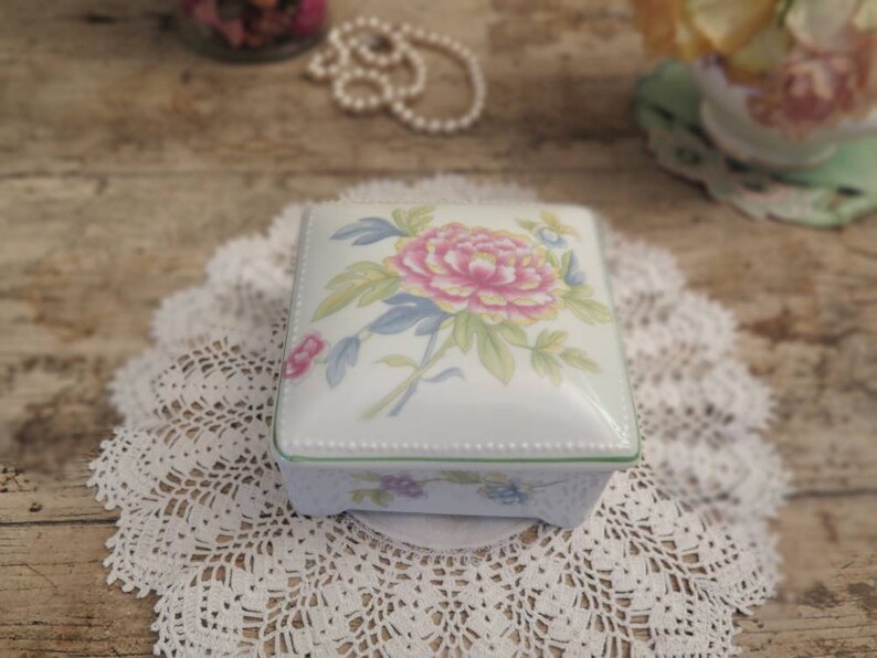 May include: A white porcelain square trinket box with a floral design. The box has a green trim and a delicate floral pattern with a pink flower in the centre.