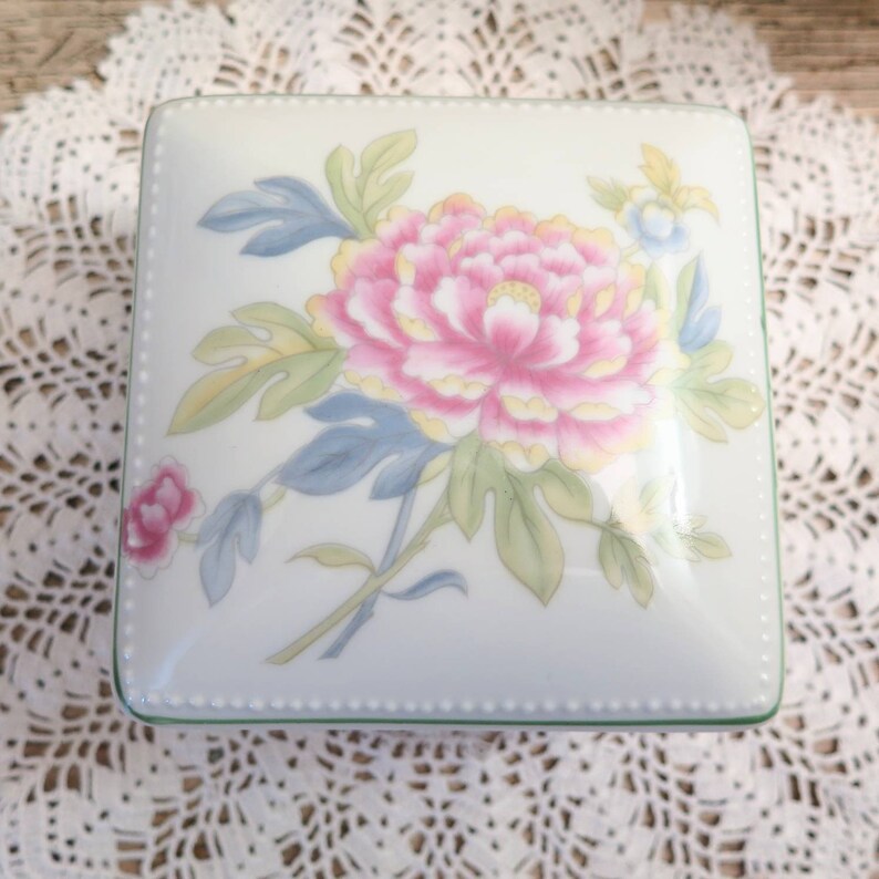 May include: A white porcelain square box with a green trim and a floral design. The design features a large pink flower with yellow accents and green leaves.