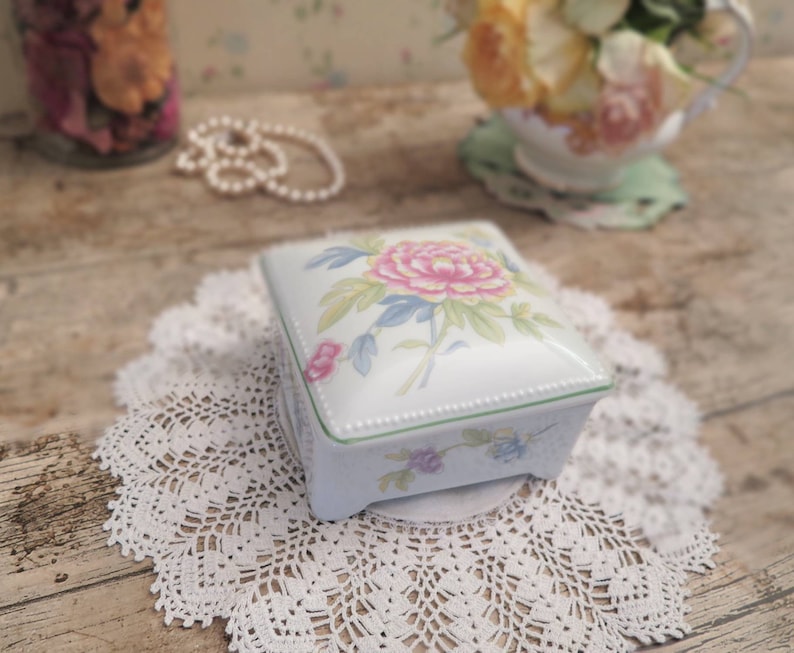 May include: A white porcelain trinket box with a floral design. The box has a green trim and a delicate floral pattern with pink, yellow, and green flowers. The box is sitting on a white crocheted doily.