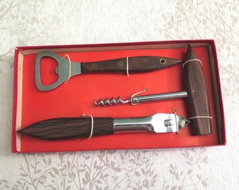 May include: A vintage bar tool set in a red box. The set includes a bottle opener, corkscrew, and a can opener, each with dark wood handles and silver-colored metal components. The tools are secured with rubber bands.