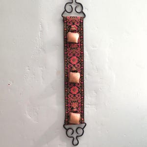 Puede incluir: Un tapiz de pared con un marco de metal negro y una larga tira de tela colorida con cuatro pequeñas campanas de color cobre. La tela tiene un patrón floral en tonos rosa, rojo y dorado.