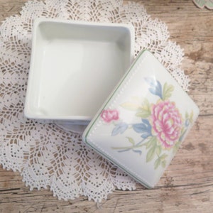 May include: A white porcelain trinket box with a floral design on the lid. The lid features a pink and yellow flower with green leaves and blue accents.