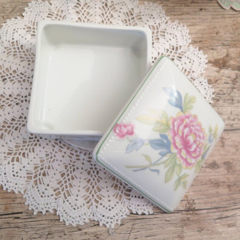 May include: A white porcelain trinket box with a floral design on the lid. The lid features a pink and yellow flower with green leaves and blue accents.