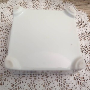 May include: White ceramic square plate with rounded edges and four raised feet on the bottom.