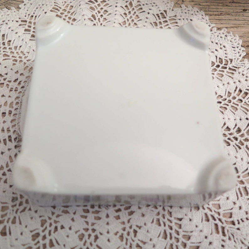 May include: White ceramic square plate with rounded edges and four raised feet on the bottom.