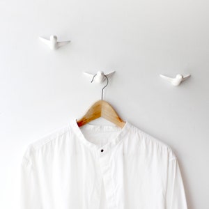 May include: A white shirt hanging on a wooden hanger with three white bird-shaped hooks on a white wall.