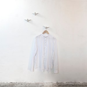 Puede incluir: Una camisa blanca con cuello colgada de una percha de madera contra una pared blanca. Tres ganchos blancos en forma de pájaro están montados en la pared.