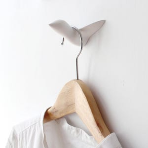 Op de afbeelding: Een witte keramische vogelvormige wandhaak met een zilveren metalen haak. Een houten hanger met een wit shirt hangt aan de haak.