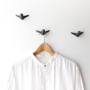 May include: A white collared shirt hanging on a wooden hanger with three black bird-shaped hooks on a white wall.