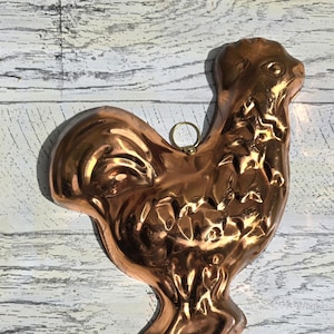 May include: A vintage copper rooster-shaped mold. The mold is a shiny, warm copper color and features detailed relief patterns. It has a small ring at the top for hanging. The rooster is standing on a wooden surface.
