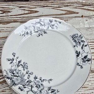 May include: A round ceramic plate with a white base and a dark gray floral pattern featuring clusters of blossoms and leaves along the rim. The plate has a smooth, glossy finish and a slightly scalloped edge with a thin metallic trim around the border.