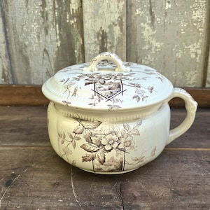 May include: Cream-colored antique chamber pot with a lid, featuring a floral and landscape pattern in brown. The pot has a handle and a matching lid with a small handle. The design includes flowers, leaves, and a scenic illustration.