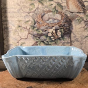 May include: A light blue ceramic rectangular dish with a textured, diamond-patterned exterior. The dish has a slightly curved rim and is set against a backdrop of a vintage-style floral print. The dish is likely used for decorative purposes.