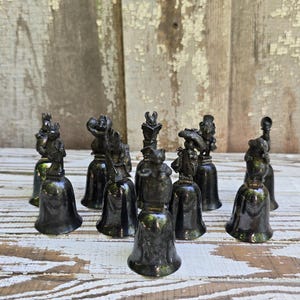 May include: A collection of nine dark metallic bells, each topped with a distinct sculpted animal figure. The bells feature various creatures, including rabbits and other small animals, standing atop smooth, flared bell-shaped bases in a dark finish.