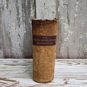 May include: An antique, cylindrical book titled "The American Elective Dispensatory" with a brown leather cover. The book has a worn appearance, with visible wear and tear. The background is a weathered, white and gray wooden surface.