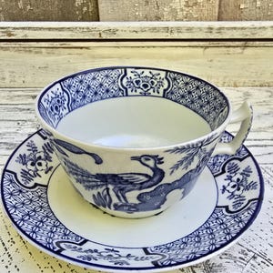 May include: A blue and white porcelain teacup and saucer set. The cup and saucer feature a detailed peacock design and intricate patterns. The set is placed on a white, distressed wooden surface, creating a vintage aesthetic.