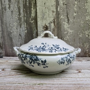 May include: A round ceramic tureen with a matching lid, featuring a white base color and a dark blue floral pattern. The piece includes two side handles and a decorative handle on the lid, with a smooth, glossy finish and intricate botanical details.