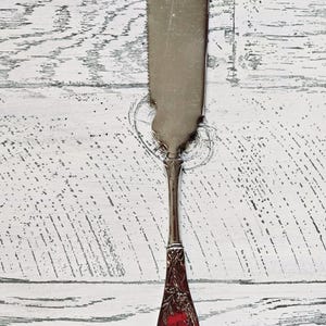 May include: A silver butter knife with a red glass handle. The handle has a floral design.