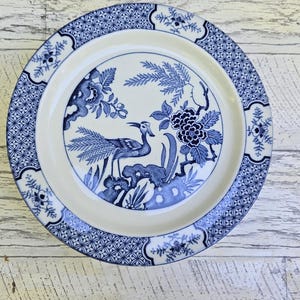 May include: A round, white and blue decorative plate with a bird and floral design. The plate features a central image of a bird with a long neck and tail, surrounded by flowers and foliage. The rim has a geometric pattern.