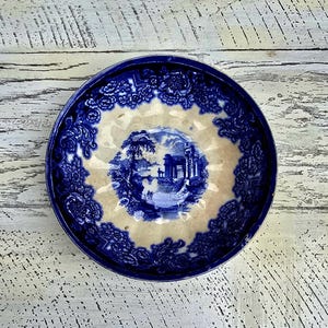 May include: A round, antique blue and white ceramic plate. The plate features a central landscape scene in blue, surrounded by a cream-colored border and a detailed blue floral pattern. The plate is set against a distressed white wooden surface.