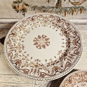 May include: A large, round, antique-style plate with a cream-colored center and a brown floral pattern. The plate has a scalloped edge and a detailed design, perfect for serving food or as a decorative piece.