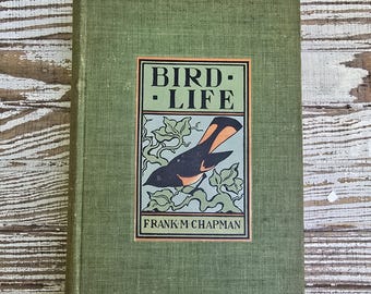 May include: A hardcover book titled Bird-Life by Frank M. Chapman with a green fabric cover. The front cover features a rectangular illustration of a black and orange bird perched on leafy branches, framed by a black border on a light green background.