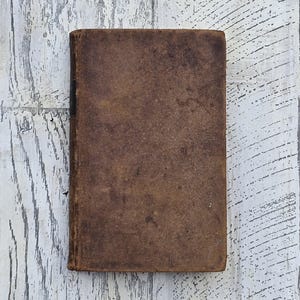 Puede incluir: Un libro cerrado, rectangular, con una cubierta de cuero marr&oacute;n desgastada. Los bordes del libro son ligeramente redondeados y la cubierta muestra signos de desgaste y envejecimiento. El fondo es una superficie de madera blanca con vetas grises.