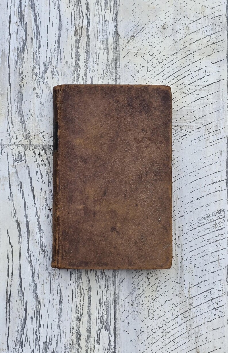 Puede incluir: Un libro cerrado, rectangular, con una cubierta de cuero marr&oacute;n desgastada. Los bordes del libro son ligeramente redondeados y la cubierta muestra signos de desgaste y envejecimiento. El fondo es una superficie de madera blanca con vetas grises.