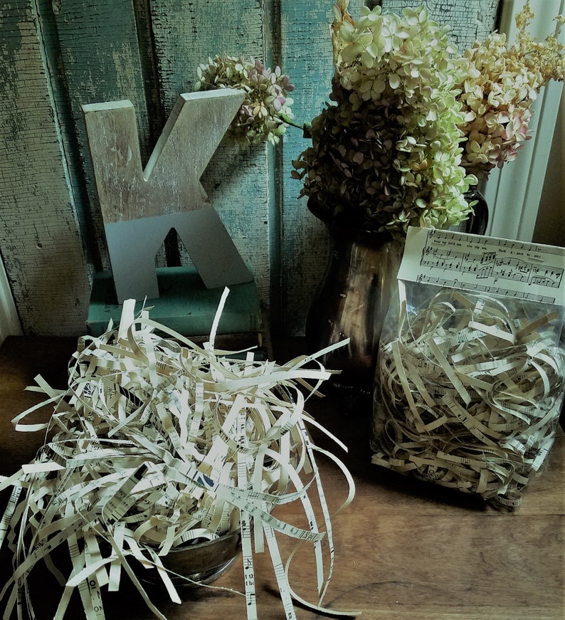 May include: Two bags of shredded sheet music, one in a clear plastic bag with a music sheet label, and the other in a brown paper bag. The bags are sitting on a wooden surface with a wooden letter K and a vase of dried flowers in the background.