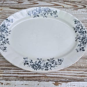 May include: An oval-shaped white ceramic serving platter featuring a scalloped edge and a decorative border of dark gray floral patterns. The design consists of detailed blossoms and leafy vines arranged symmetrically around the rim of the dish.