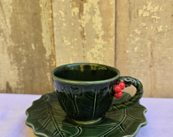 May include: A green ceramic teacup and saucer set with a holly leaf design. The cup has a handle shaped like a holly sprig with red berries.
