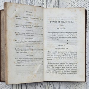 Puede incluir: Libro antiguo abierto con p&aacute;ginas envejecidas y amarillentas. La p&aacute;gina izquierda muestra una tabla de contenidos, mientras que la p&aacute;gina derecha muestra el t&iacute;tulo "The Power of Religion, &c" y el comienzo del Cap&iacute;tulo 1. El libro tiene una cubierta de cuero marr&oacute;n.