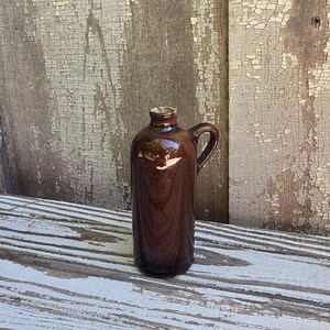 May include: A small, cylindrical brown ceramic jug with a glossy finish, featuring a narrow neck, a single curved handle on the side, and a smooth, rounded body. The vessel has a simple, functional design with a uniform dark brown glaze throughout.